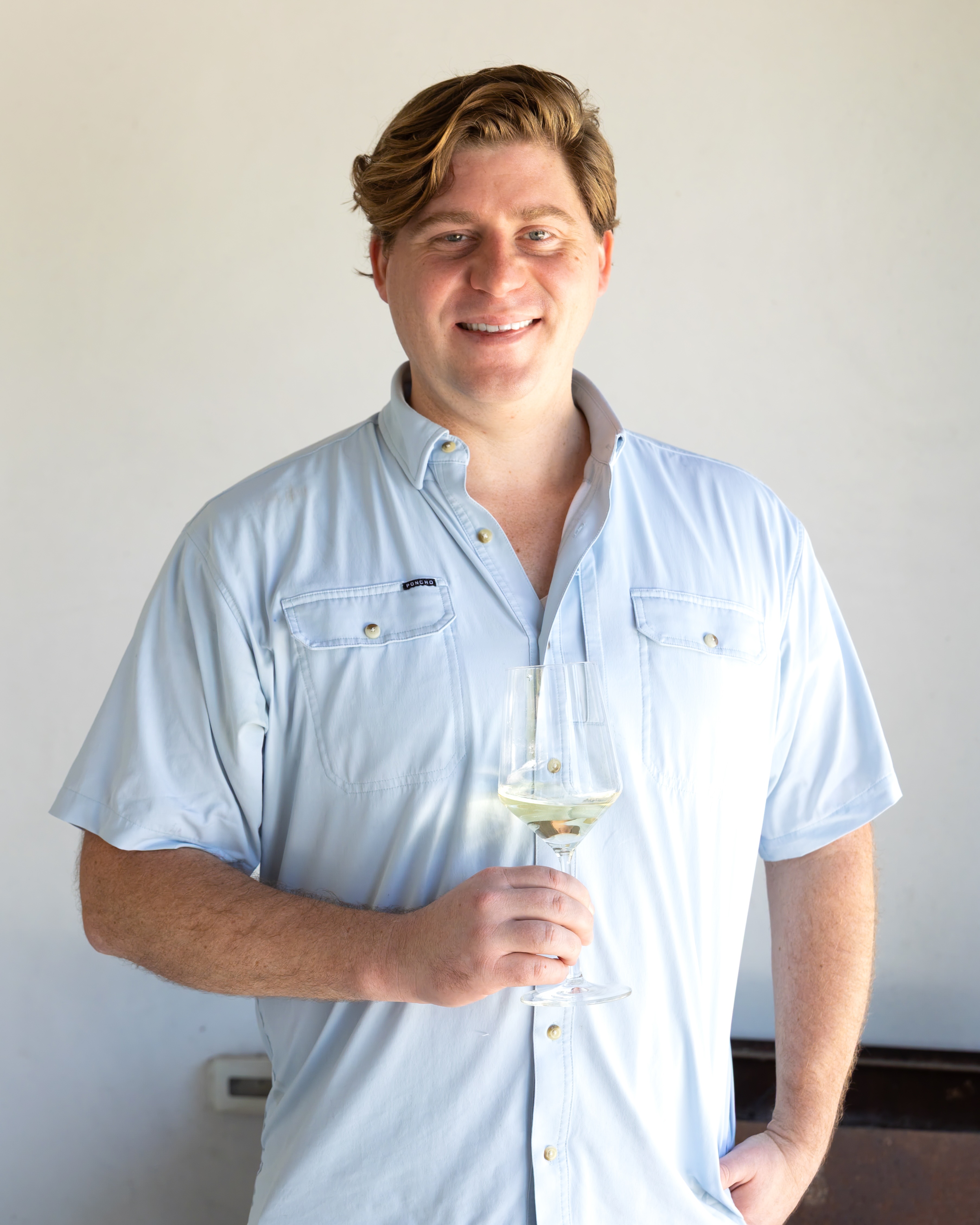 Master Sommelier Jack Mason wearing a light blue short-sleeve button-up shirt holding a stemmed wine glass with white wine.