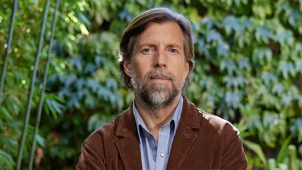 Bearded man with medium-length brown hair and mustache, wearing a blue collared shirt and brown jacket, standing outdoors with green foliage background.