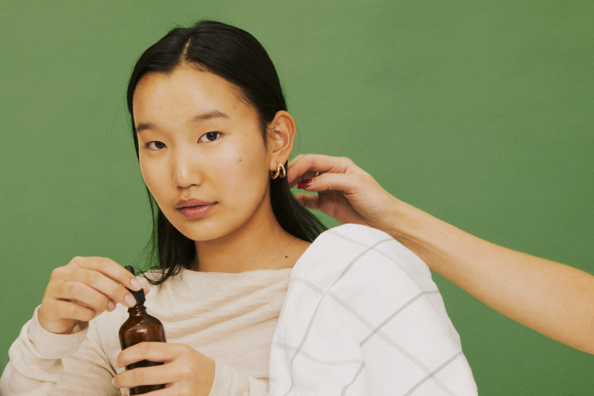Model holding a serum bottle against a green backdrop while a stylist adjusts her hair