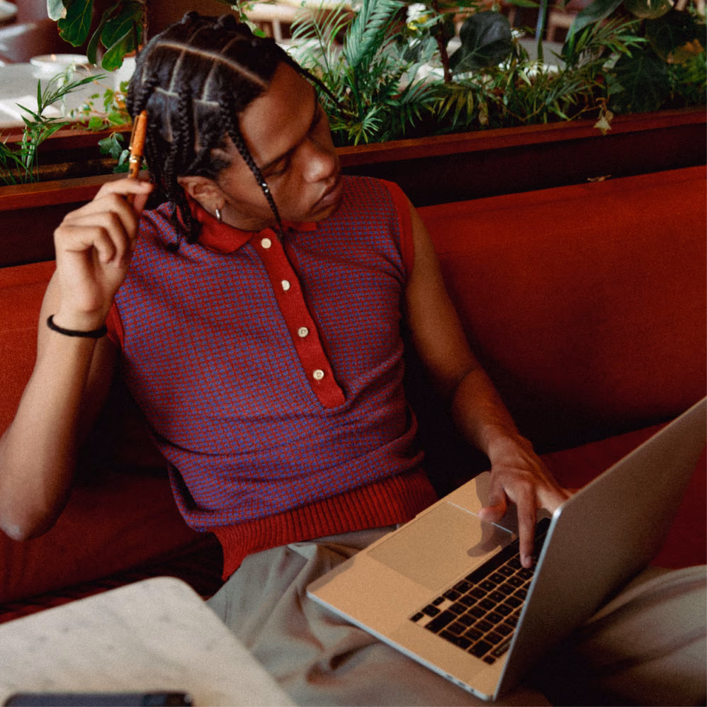 Copywriter working on a laptop on a red velvet sofa, pen held to their braided hair