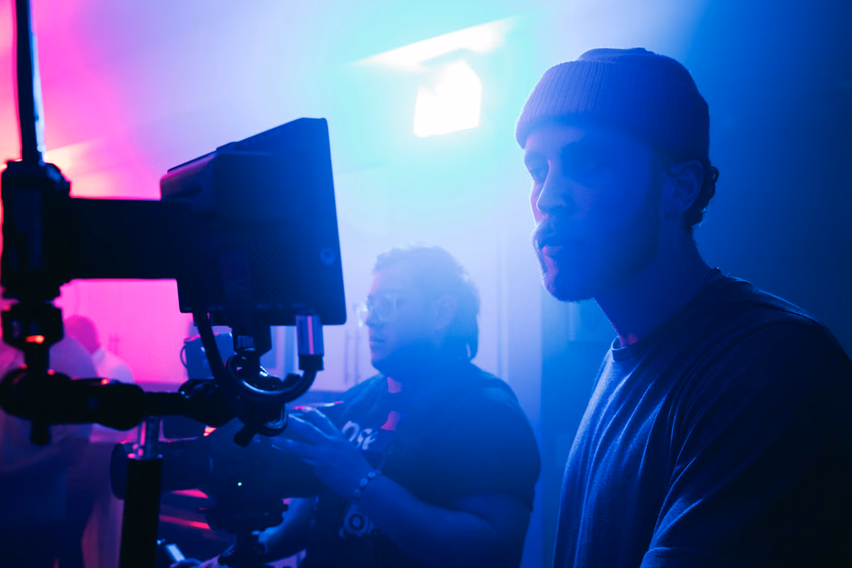 Crew member beside a camera monitor on a set lit with pink and blue light