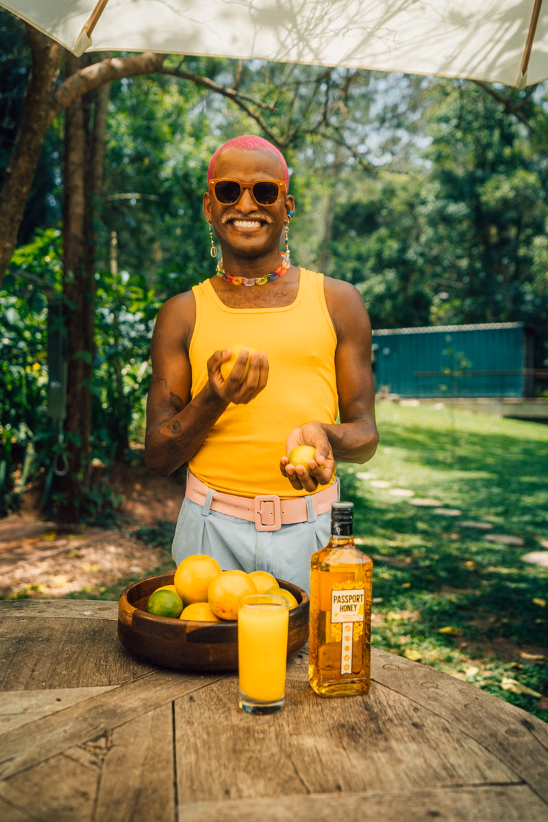Creator in a yellow vest holding fruit beside a bottle and glass of juice on a garden table