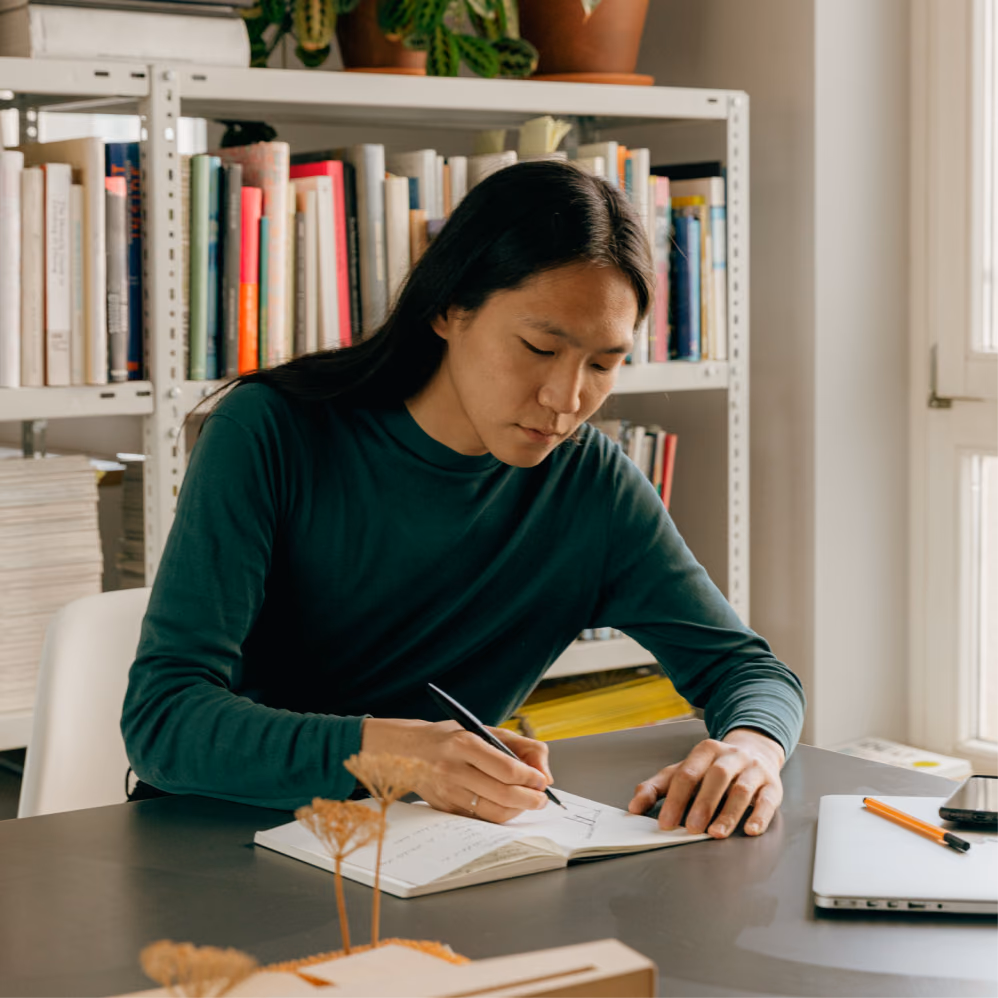 Writer making notes in a notebook at a desk in front of a full bookshelf