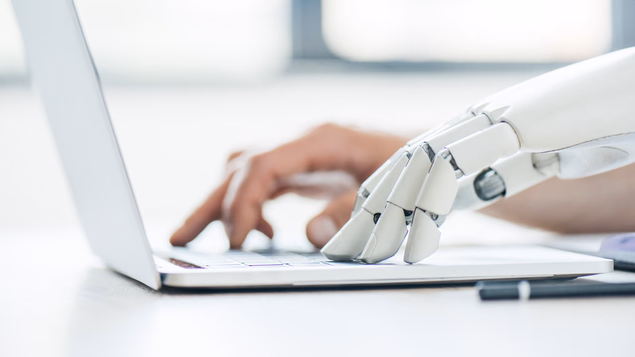 A robotic hand and a human hand typing together on a laptop keyboard.