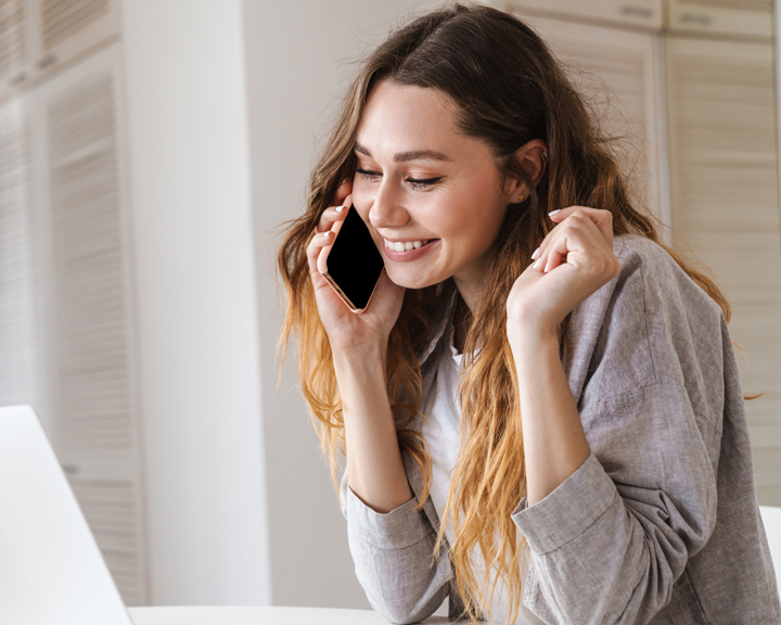A smiling woman talking on a smartphone while sitting at a desk with a laptop.