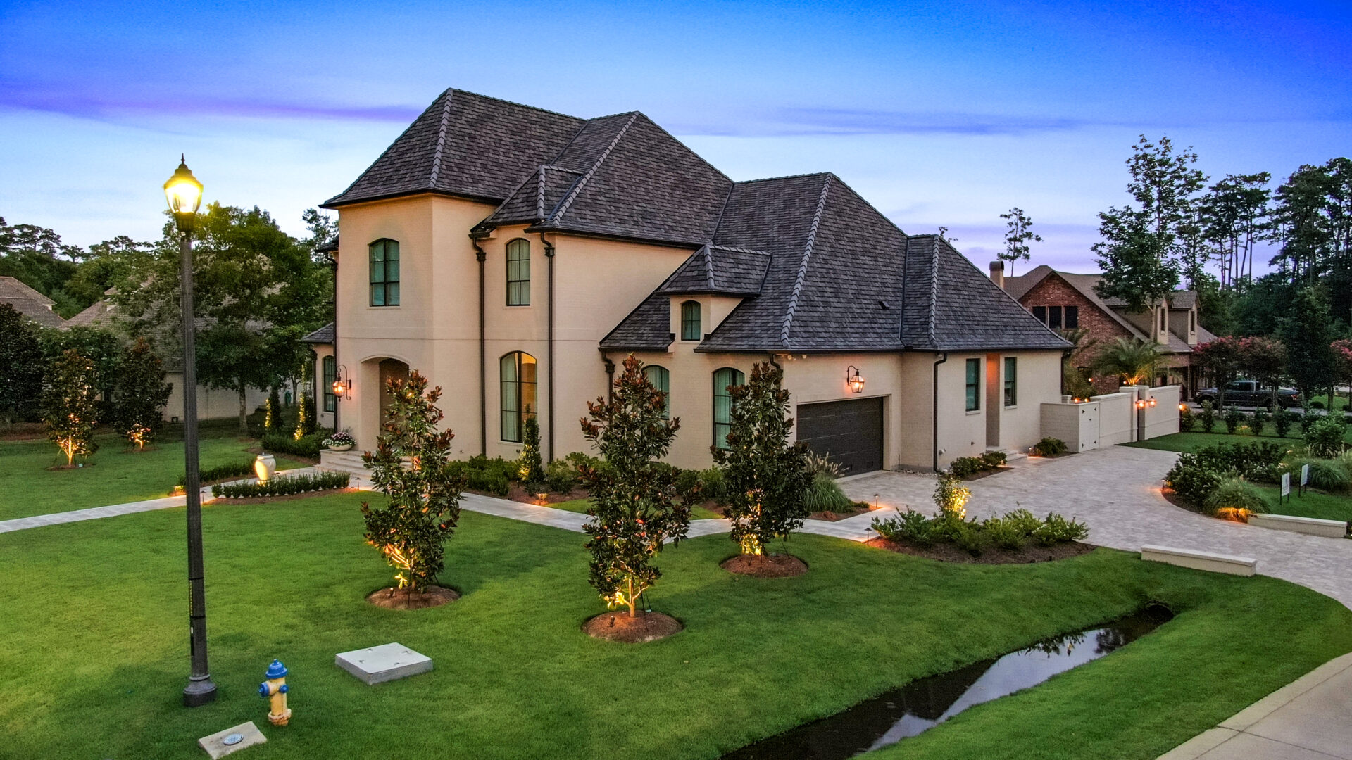 A large two-story beige house with a dark shingled roof, a driveway leading to a garage, neatly landscaped lawn with illuminated small trees, a street lamp, and a narrow water drainage ditch.