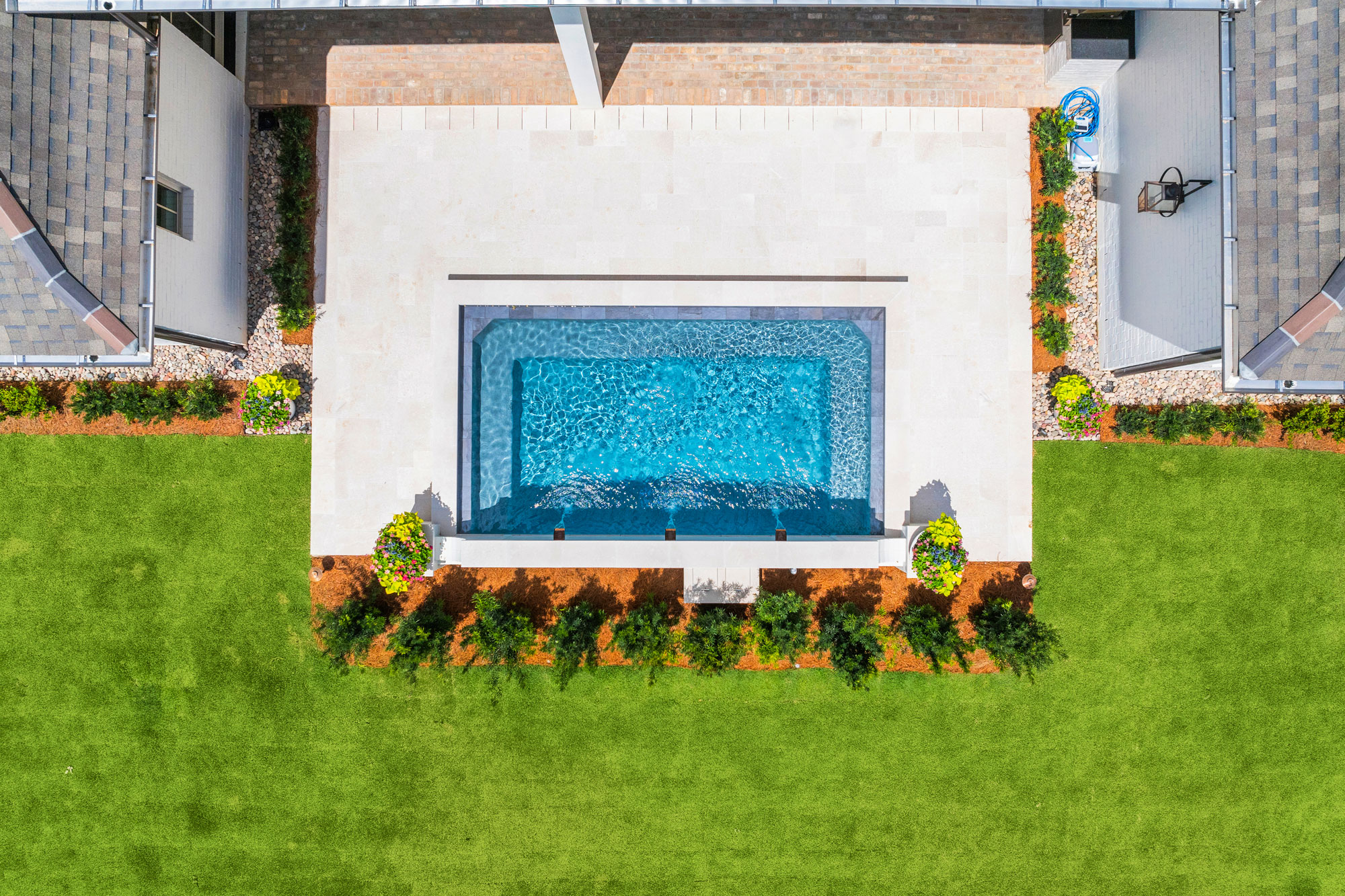 Aerial view of a rectangular backyard pool with blue water, surrounded by white stone patio and green lawn.