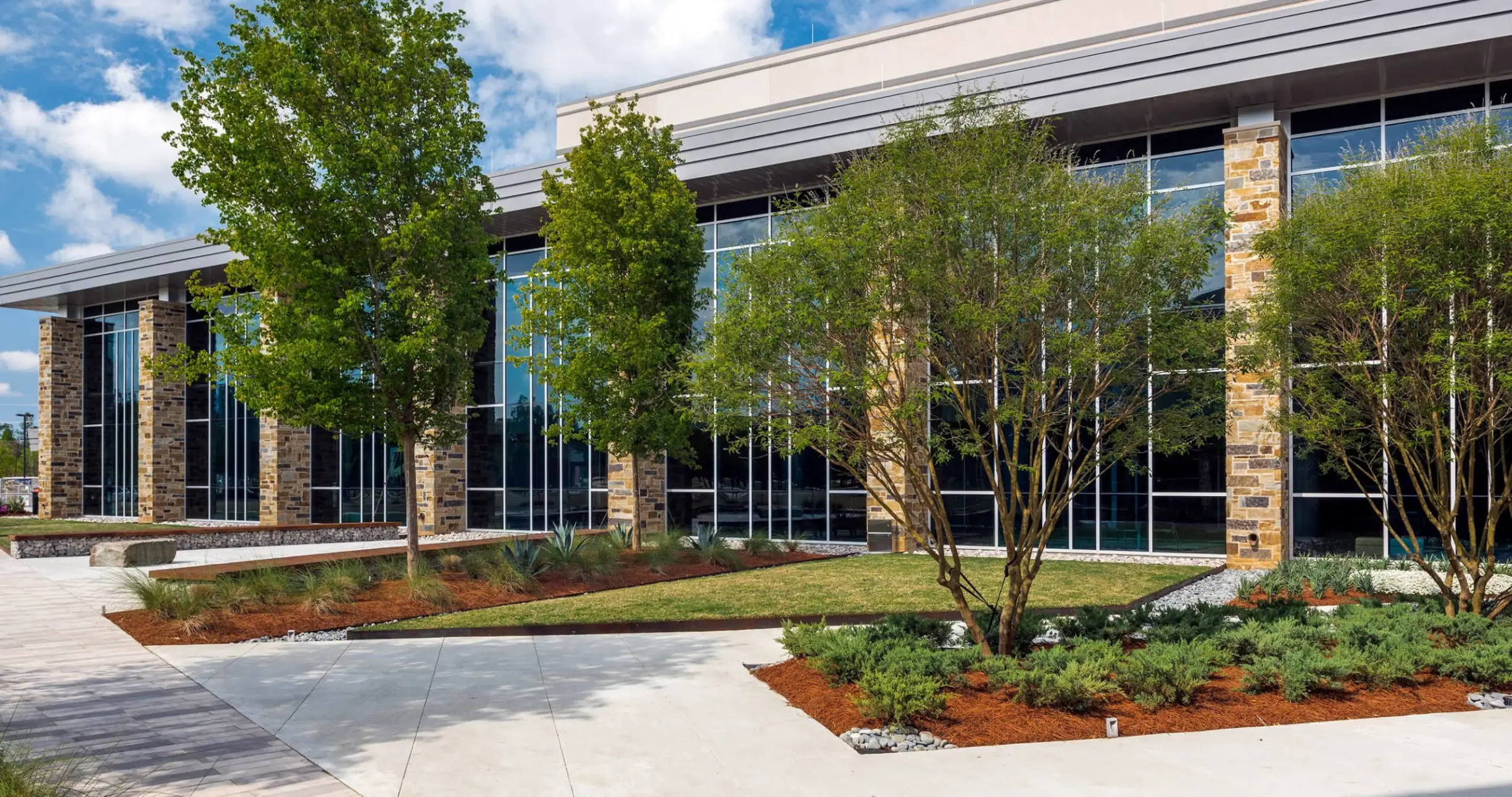 Modern building with large glass windows, stone columns, and landscaped greenery under a blue sky.