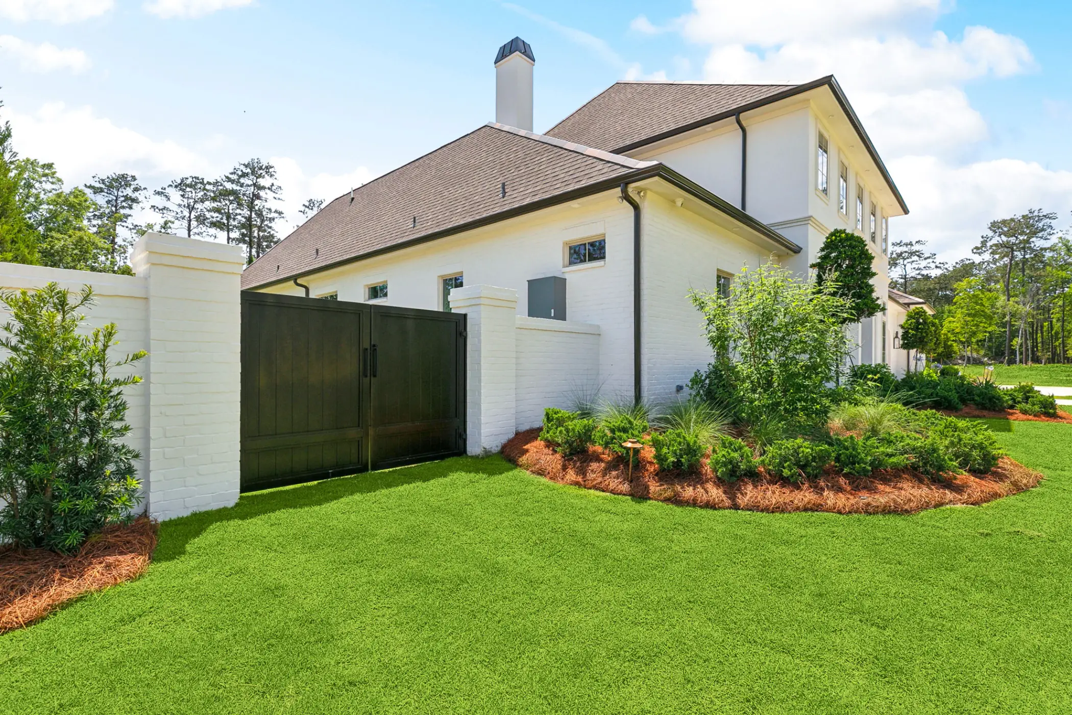 Side view of a white brick house with brown roof, black gate, and landscaped green lawn.