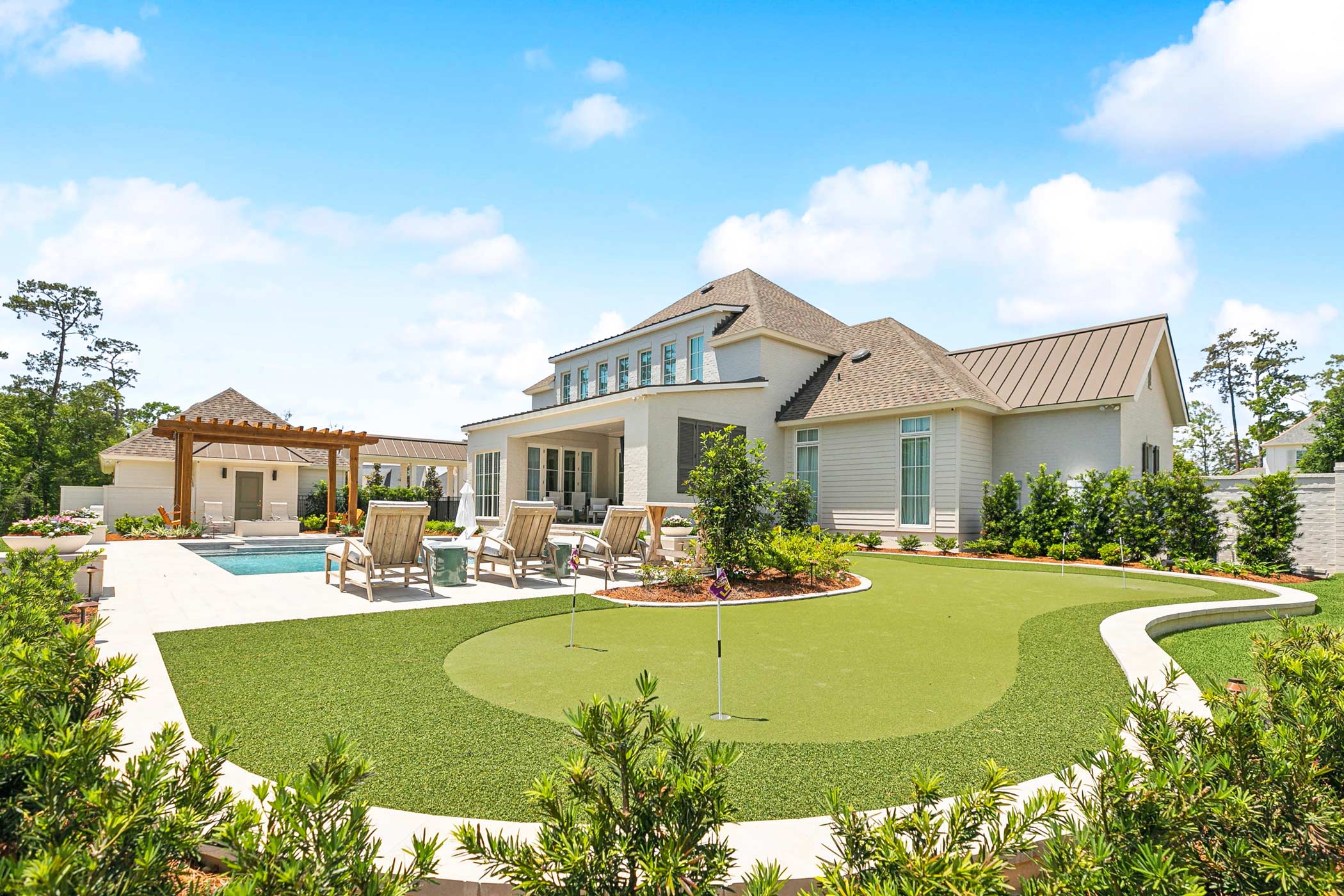 Modern white house with a large backyard featuring a swimming pool, lounge chairs under a wooden pergola, a covered patio, and a putting green surrounded by landscaping.