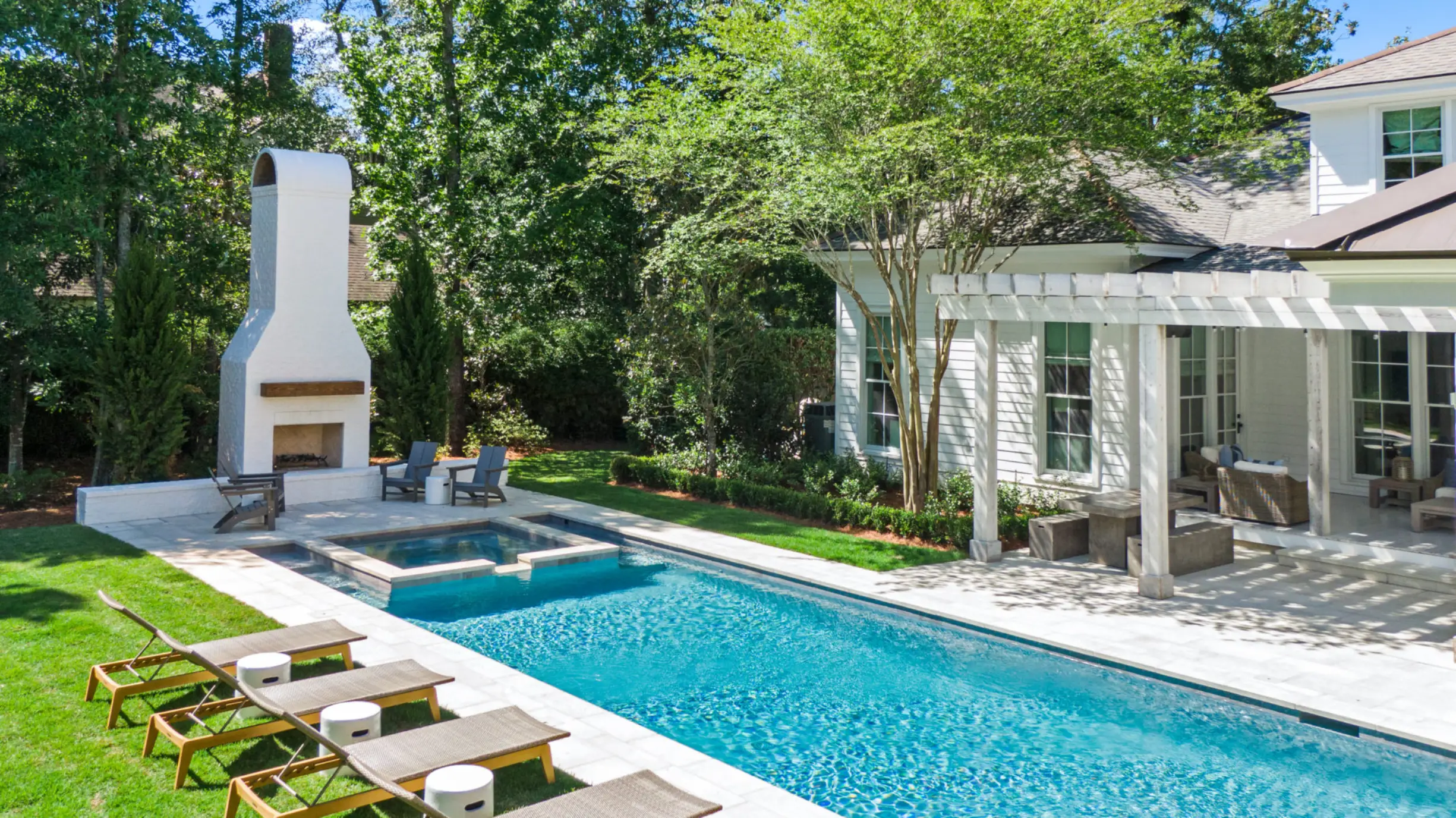 Tchefuncte Estates outdoor pool area by Smoketree Landscape and Wetstone Custom Pools with a rectangular blue pool, adjacent spa, four lounge chairs with side tables on grass, white fireplace with seating, and a white house with a patio and pergola.