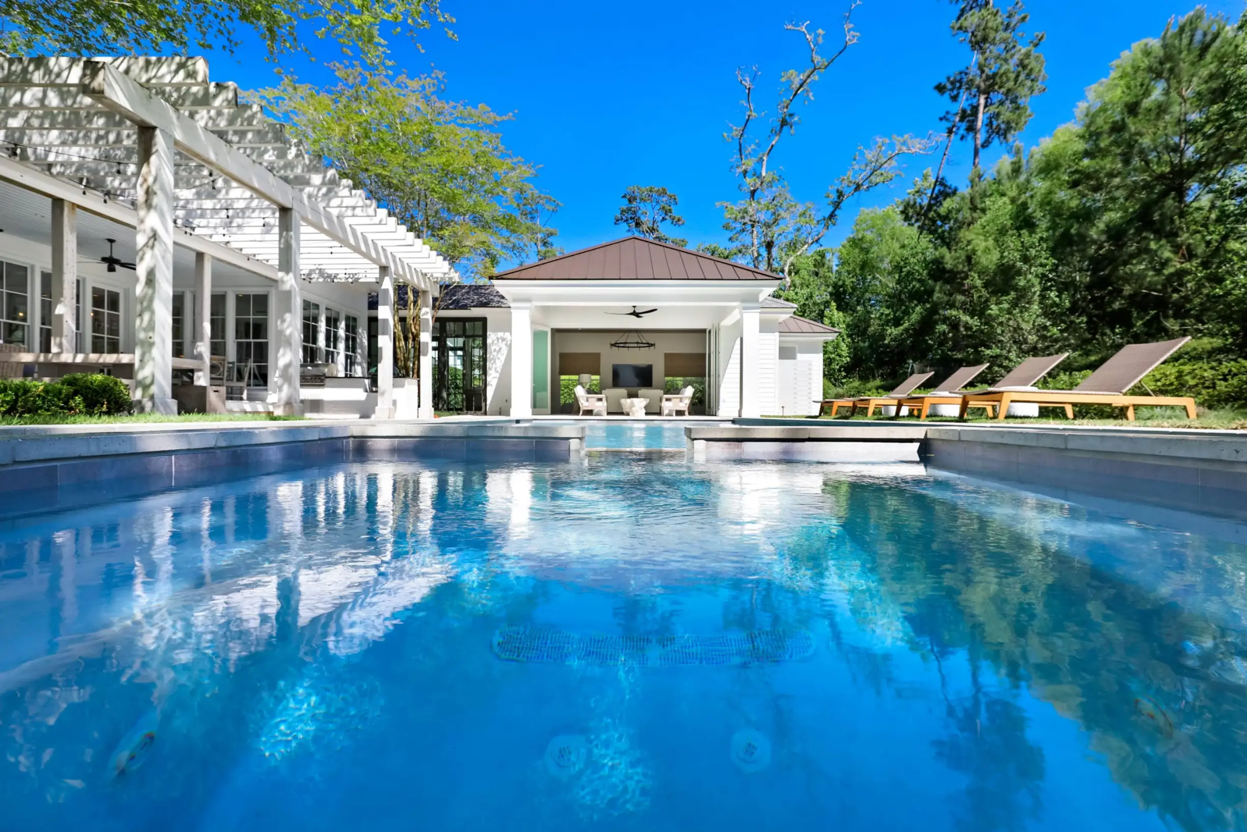Clear blue swimming pool in front of a white house with a covered patio and pergola, surrounded by green trees and with lounge chairs on the right side.