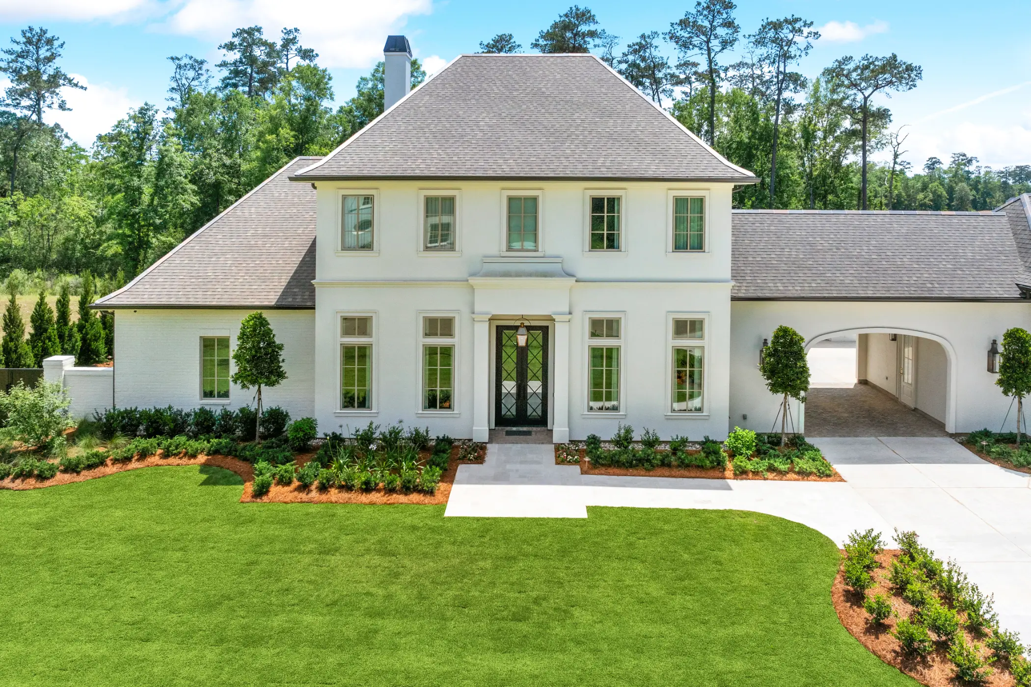 Two-story white house with gray shingled roof, multiple windows, black front door, manicured lawn, garden beds, a driveway, and an arched carport on the right, surrounded by tall trees.