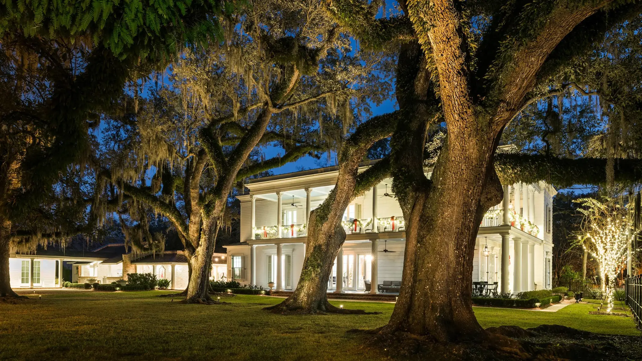 Large white two-story house with illuminated porches and columns, decorated with wreaths and lights, surrounded by big trees with hanging moss at dusk.