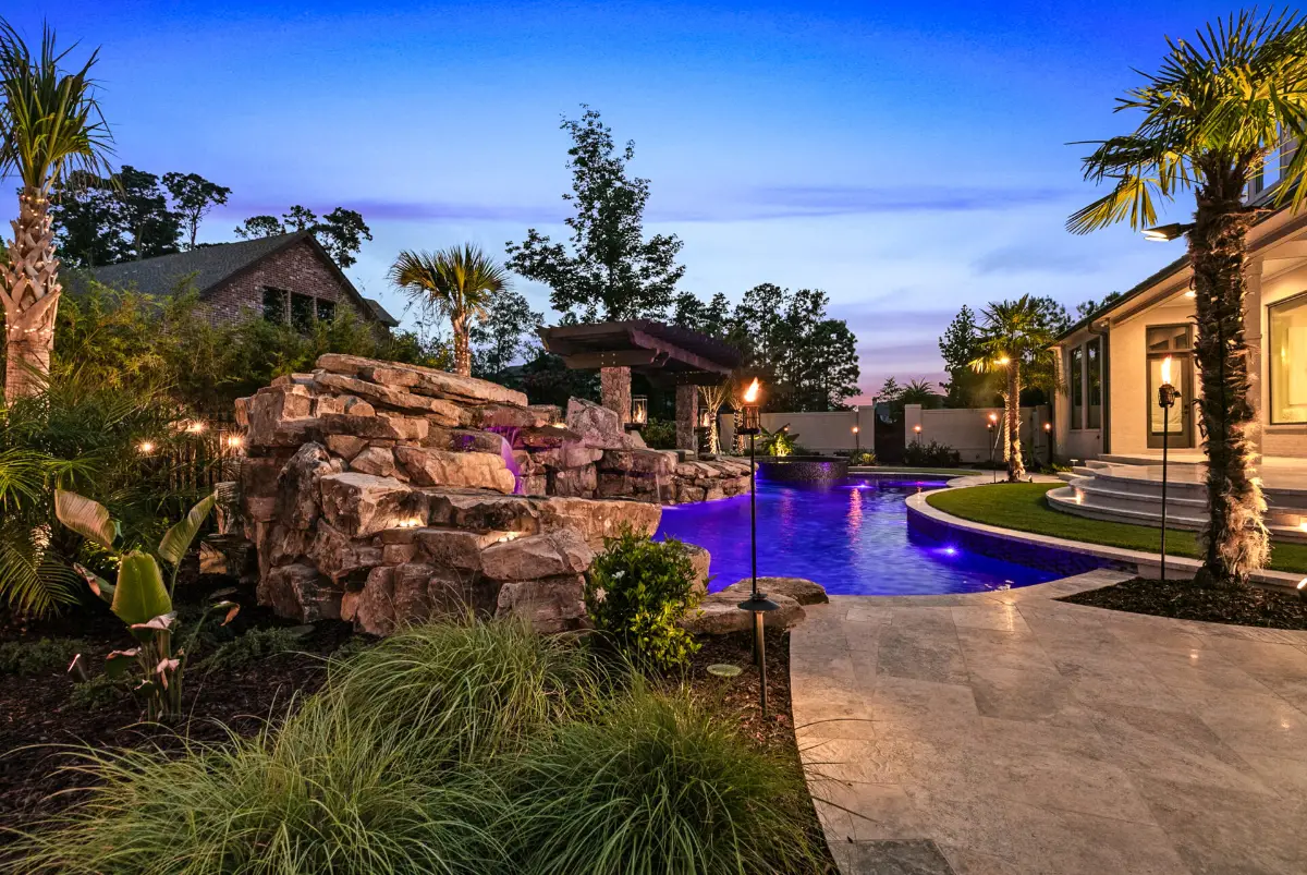 Luxurious outdoor pool area at dusk with a waterfall feature made of rocks, surrounded by palm trees and lush greenery, illuminated by soft purple pool lights and tiki torches, adjacent to a modern house with curved steps.