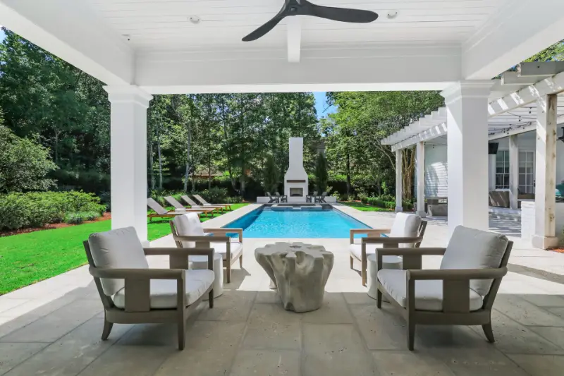 Covered patio with four cushioned armchairs arranged around a white stone coffee table, overlooking a rectangular swimming pool with a white outdoor fireplace at the far end, surrounded by green lawn and trees.
