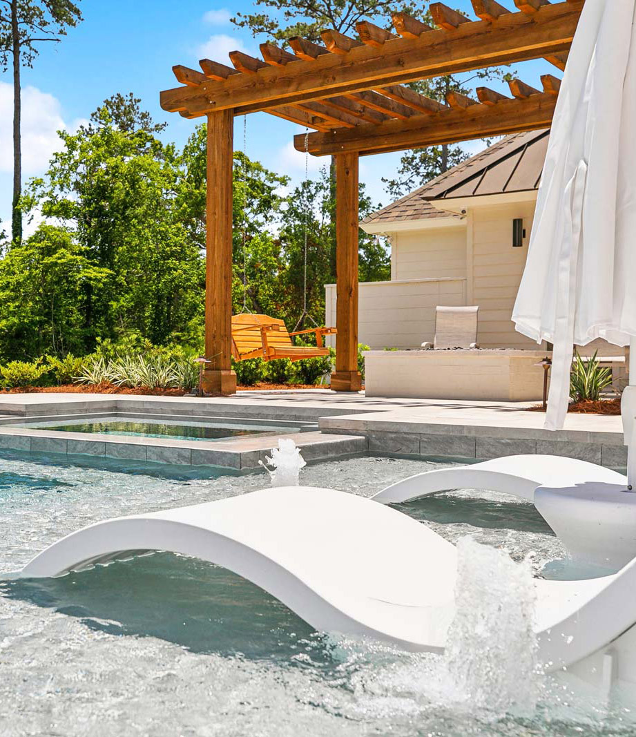 Outdoor pool area with a shallow lounging section featuring white curved lounge chairs partially submerged in water, a closed white umbrella, and two small water fountains, bordered by a hot tub. In the background is a wooden pergola with a hanging wooden swing and greenery under a bright blue sky.