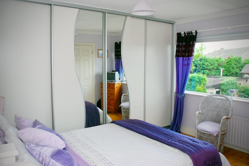 Bedroom with a bed covered in white and purple bedding, purple pillows, a wicker chair with a cushion near a window with purple and black curtains, and a white wardrobe with mirrored sliding doors reflecting a dresser and part of the room.