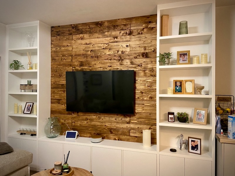 Living room wall with wooden paneling behind a mounted flat-screen TV, flanked by white built-in shelves holding framed photos, plants, candles, and decorative items, with white storage cabinets below and part of a beige sofa visible on the left.