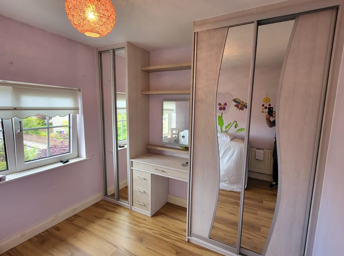 Bedroom corner with light wood laminate floor and lavender walls, featuring a white-framed window with a roller blind to the left. A built-in light wood wardrobe with mirrored sliding doors is to the right, next to a small built-in desk with drawers, open shelving, and a mirror above it. A round orange ceiling light fixture hangs above. Reflections in the mirrors show part of a bed, a person taking the photo, a wall radiator with a white towel, and wall decals of butterflies and flowers.