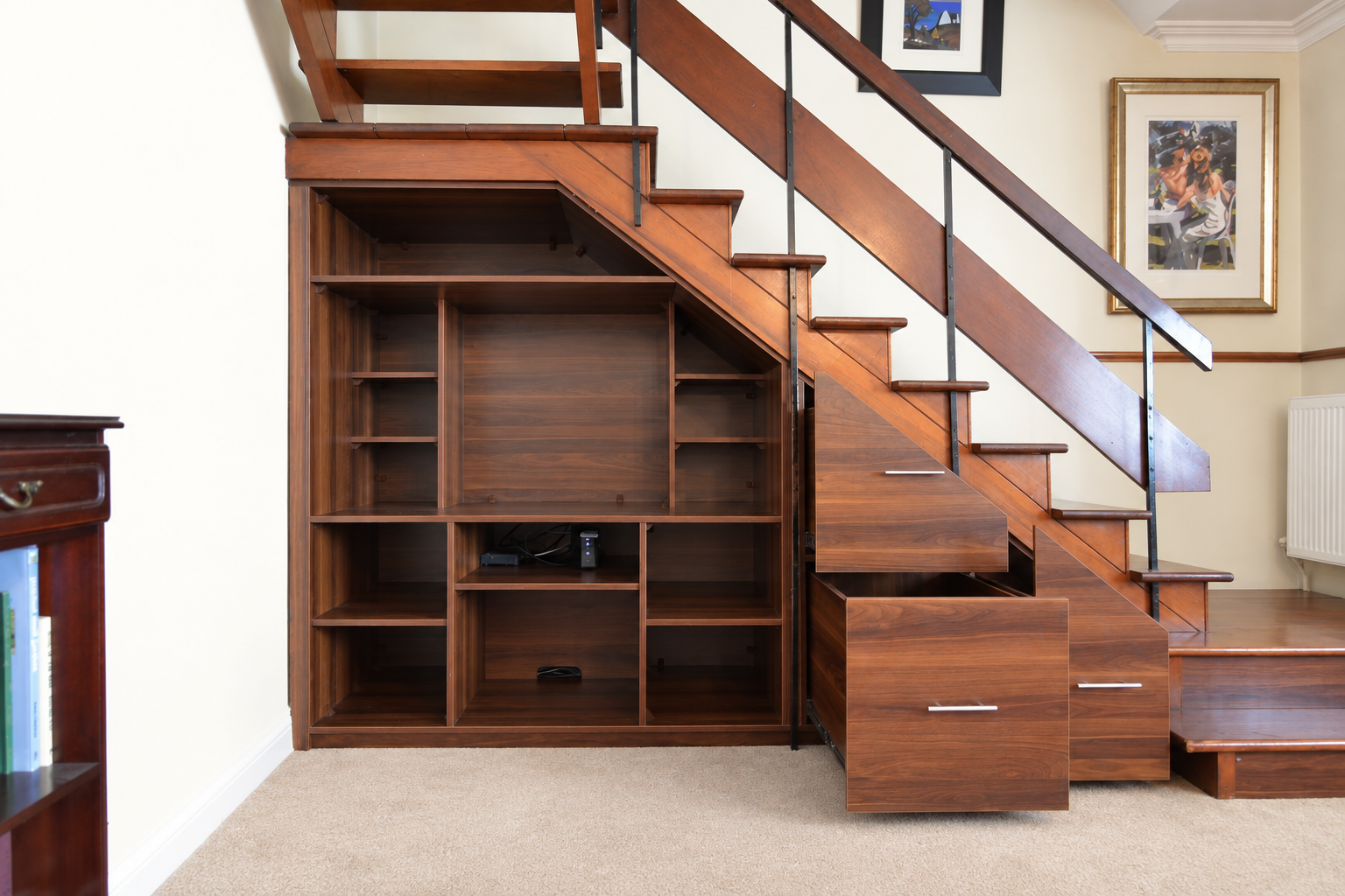 Wooden built-in storage unit with open shelves and drawers under a wooden staircase with metal railings in a carpeted room with framed artwork on the walls.
