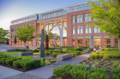 Modern three-story brick building with large windows and a glass atrium entrance, surrounded by landscaped gardens with shrubs, grass, trees, stone benches, and tall decorative stone sculptures under a clear sky at dusk.
