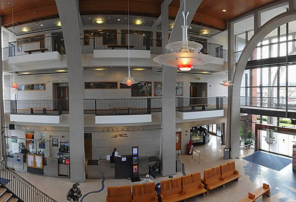 Interior view of a multi-level public building lobby with large concrete pillars, a high wooden ceiling, hanging lights, a glass entrance wall, wooden benches, and people walking or sitting.