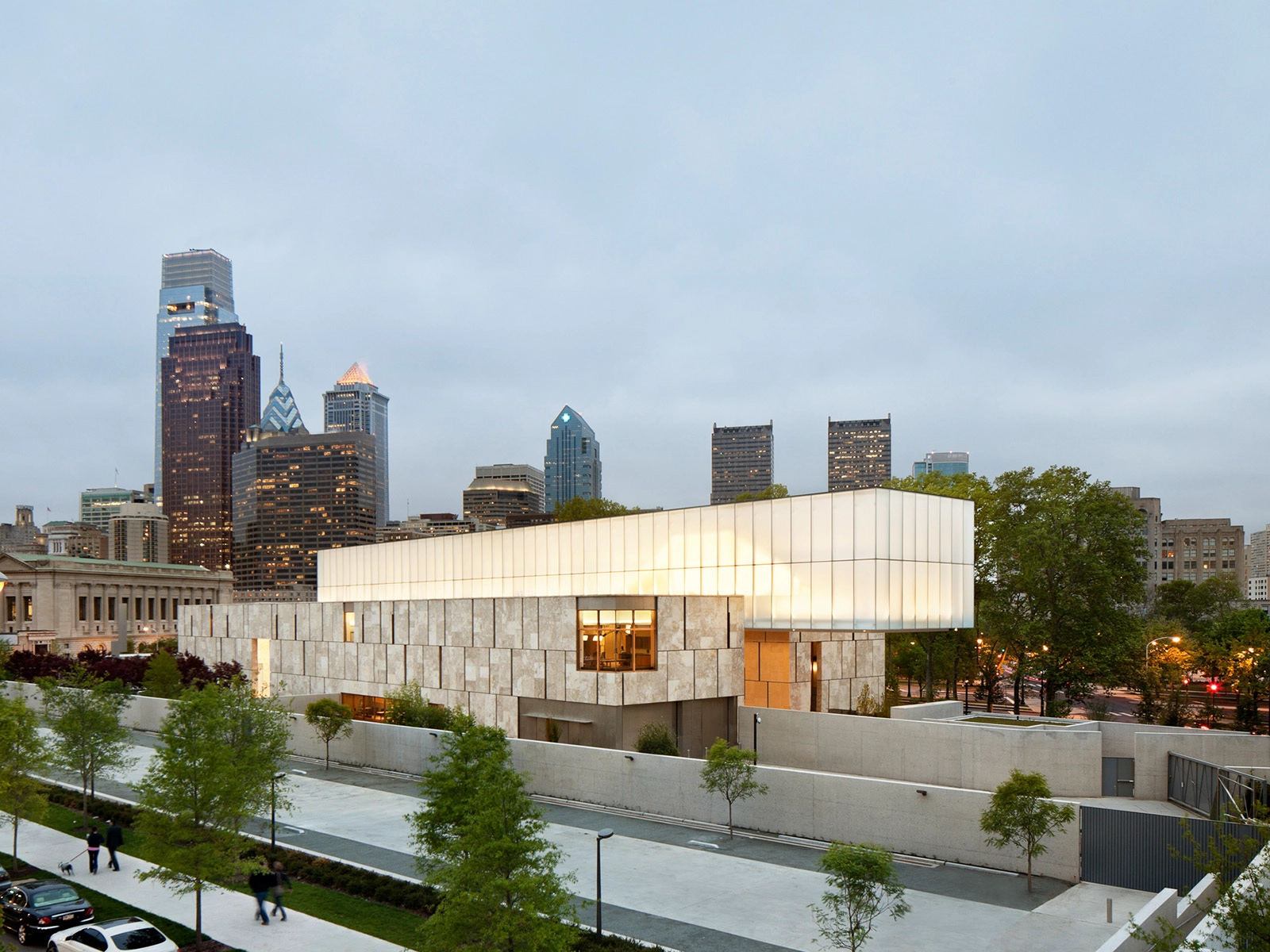 Modern building with a glowing white upper section and a stone facade lower section, surrounded by trees and walkways, with a city skyline in the background under a cloudy sky.