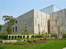 Modern building with large rectangular beige stone panels, flat roof, and glass section on top, surrounded by green lawn, shrubs, and small trees under a clear blue sky.