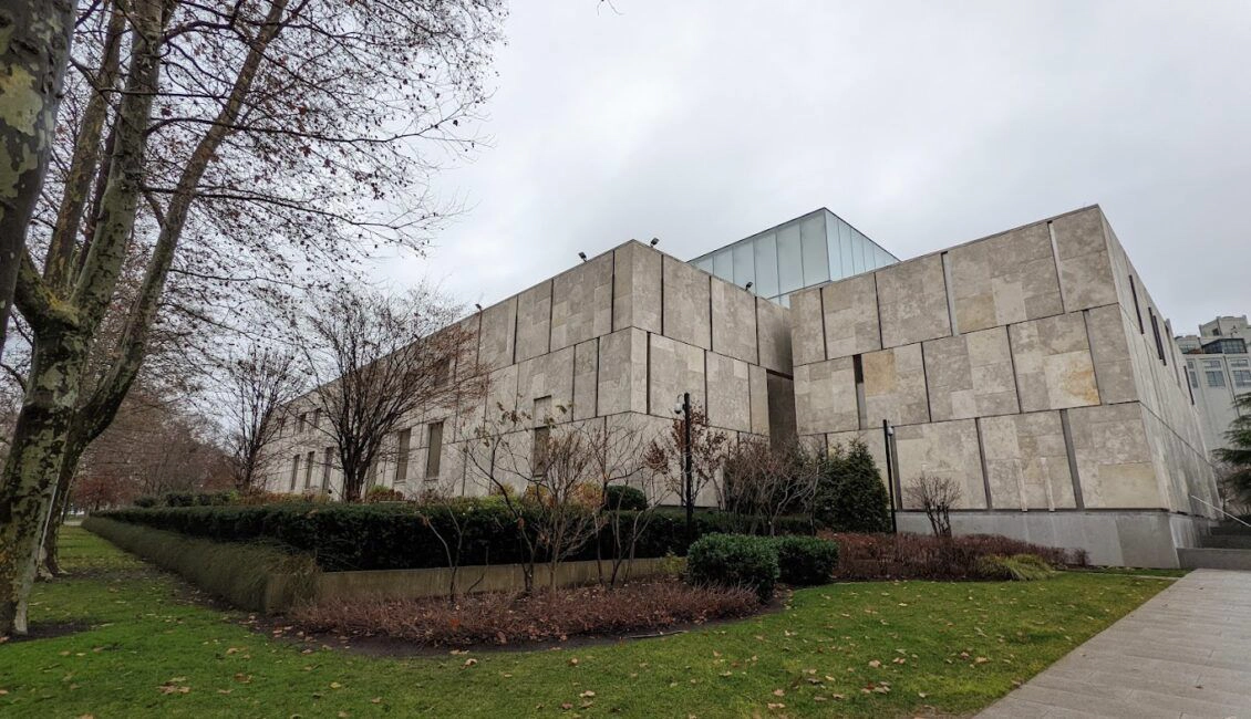 Modern concrete building with rectangular stone panels and tall, bare trees surrounding it on a grass lawn under an overcast sky.