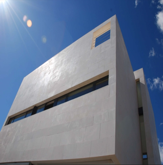 Modern white building with a smooth facade and narrow horizontal windows against a clear blue sky with sun glare in the top left corner.