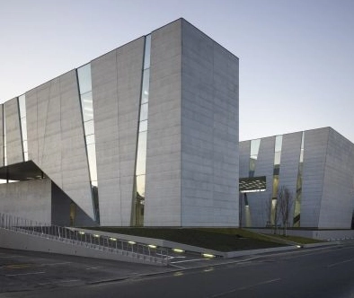 Modern architectural building with large angular concrete walls and vertical glass strips, set against a clear sky.