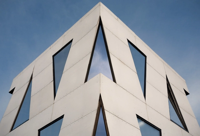 Upward view of a modern building corner with white angular panels and large, irregularly shaped glass windows reflecting the blue sky.