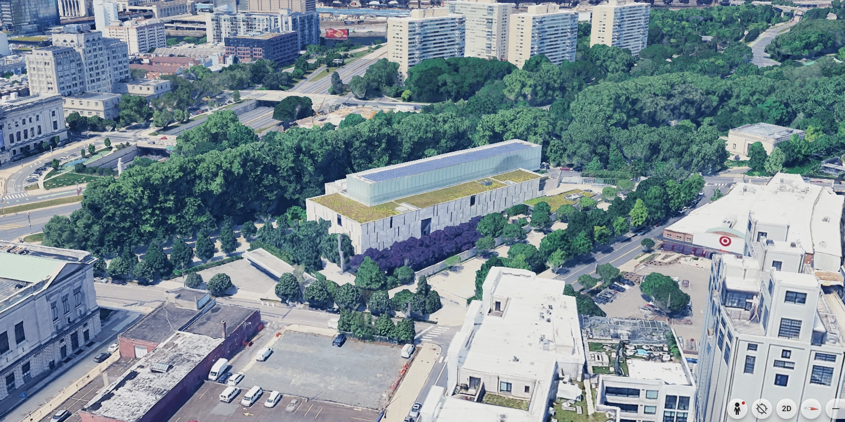 Aerial view of a modern rectangular building with a green roof and a glass upper structure, surrounded by dense trees and urban city buildings, with a parking lot visible in the foreground.