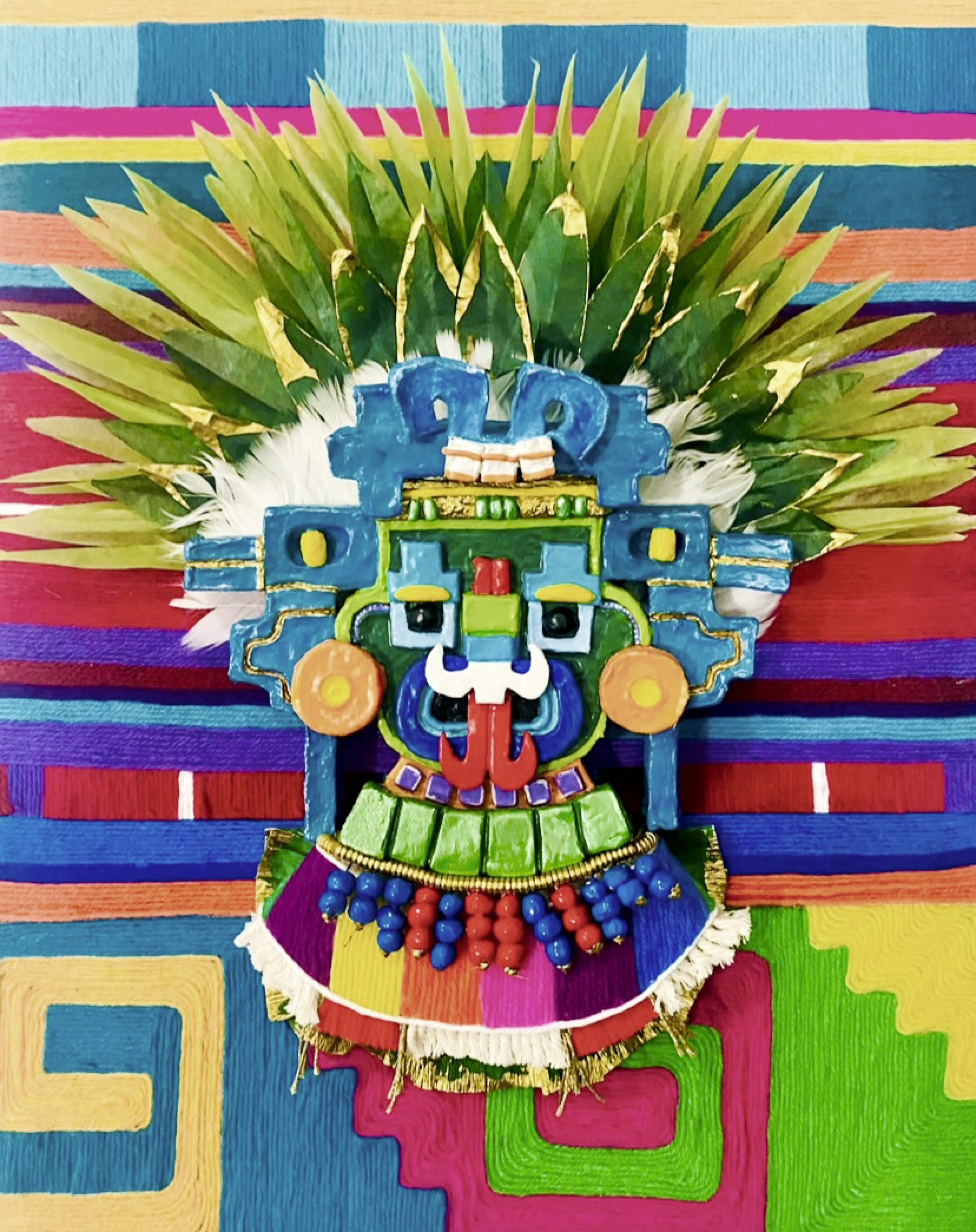 Colorful handcrafted mask with bold geometric shapes, blue and green face, red tongue, orange circular ear ornaments, and green feathers extending from the top, set against a vibrant patterned fabric background with stripes and squares in colors like red, blue, yellow, purple, and green.
