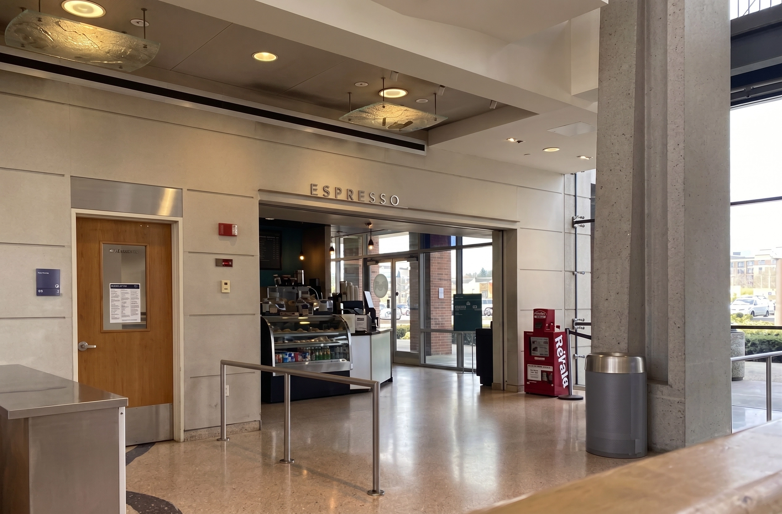 Interior view of a modern coffee shop labeled 'ESPRESSO' above the entrance with a display case of pastries and beverages inside, a wooden door to the left, and a large concrete pillar with a trash can to the right near glass windows showing a parking lot outside.