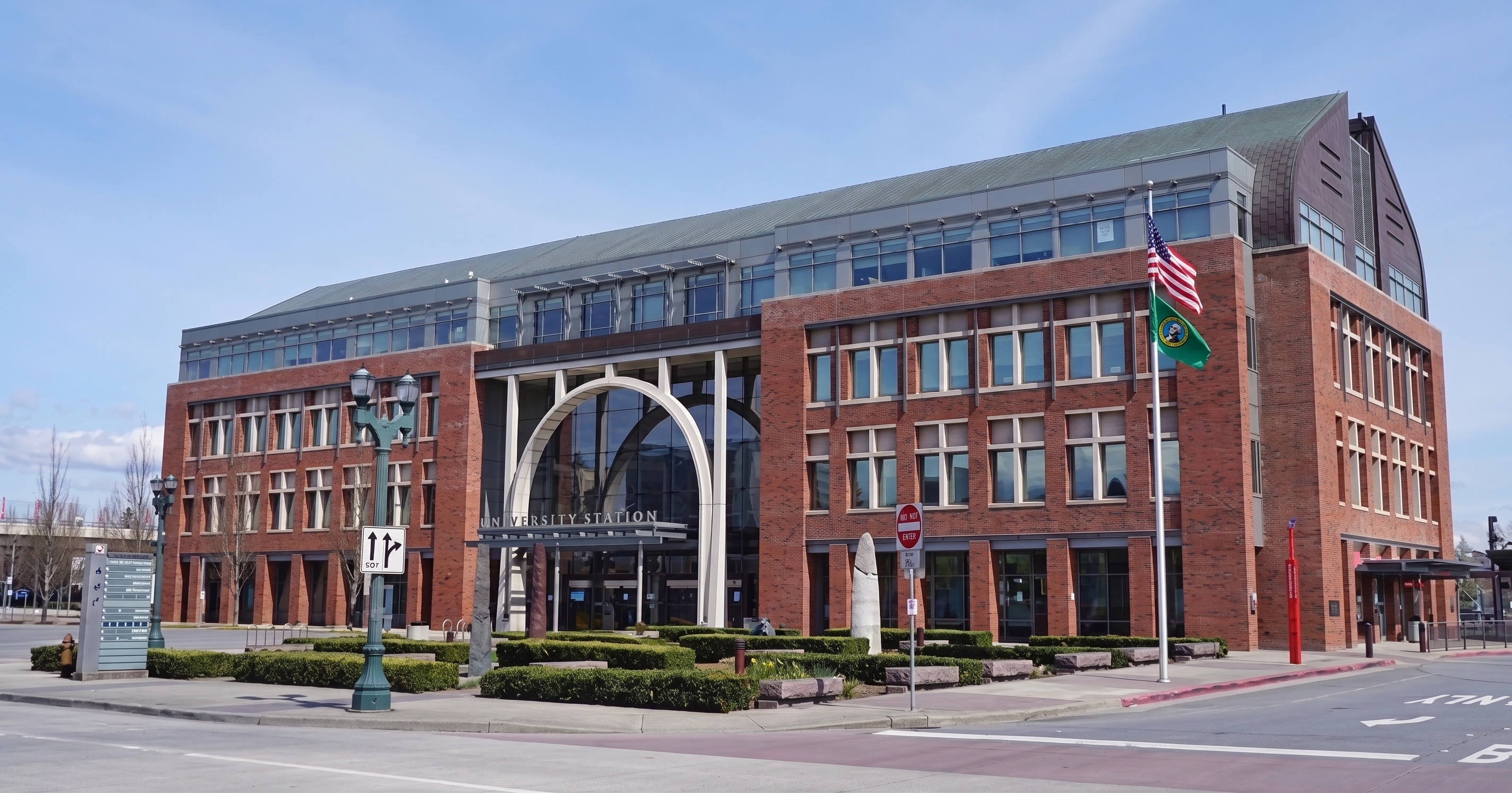 Red brick University Station building with large arched glass entrance, multiple windows, two flagpoles with the American and Washington state flags, street lamps, and trimmed hedges along the sidewalk on a clear day.