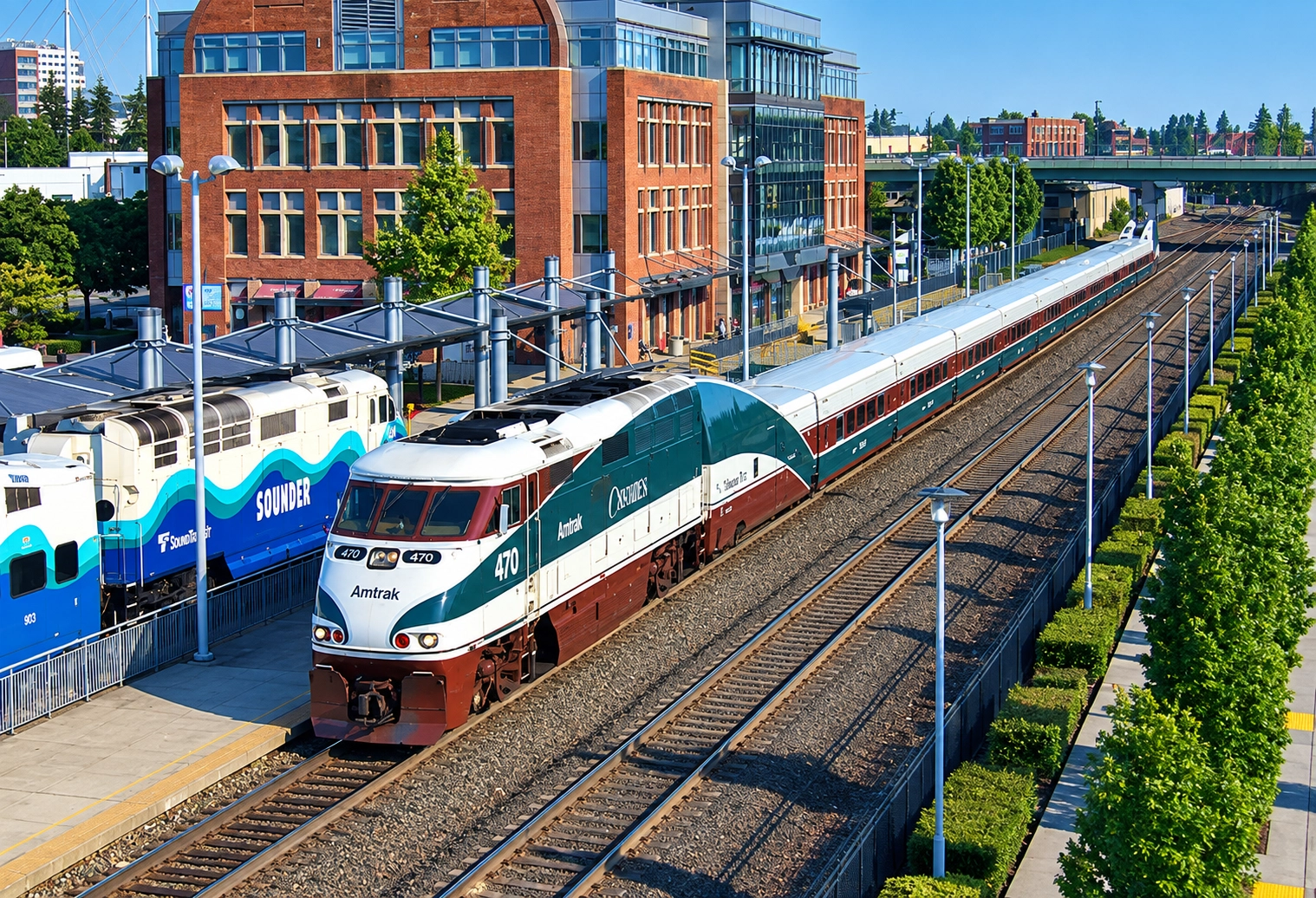 Amtrak Cascades passenger train number 470 pulled by a green, white, and maroon locomotive parked at a station platform, with a Sounder commuter train next to it and a red brick building in the background under a clear blue sky.