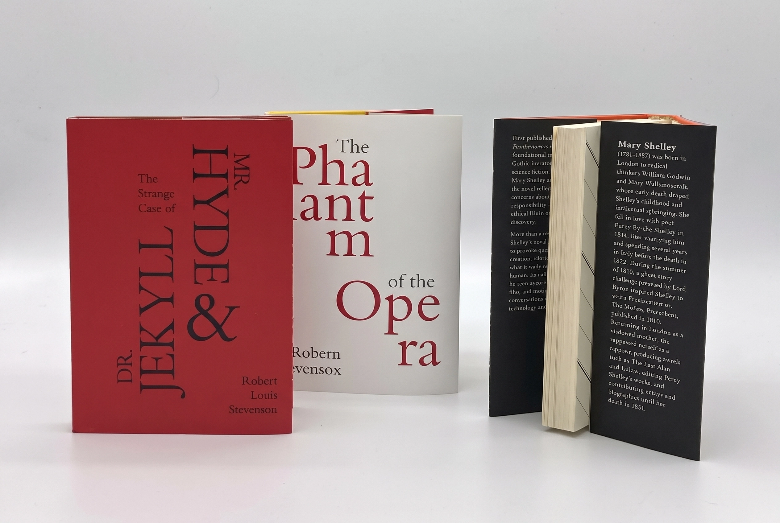 Three books standing upright on a white surface: on the left, a closed red book titled 'The Strange Case of Dr. Jekyll & Mr. Hyde' by Robert Louis Stevenson; in the center, a closed white book titled 'The Phantom of the Opera' by Robert Stevenson; on the right, an open book revealing a black inside cover page with text about Mary Shelley.