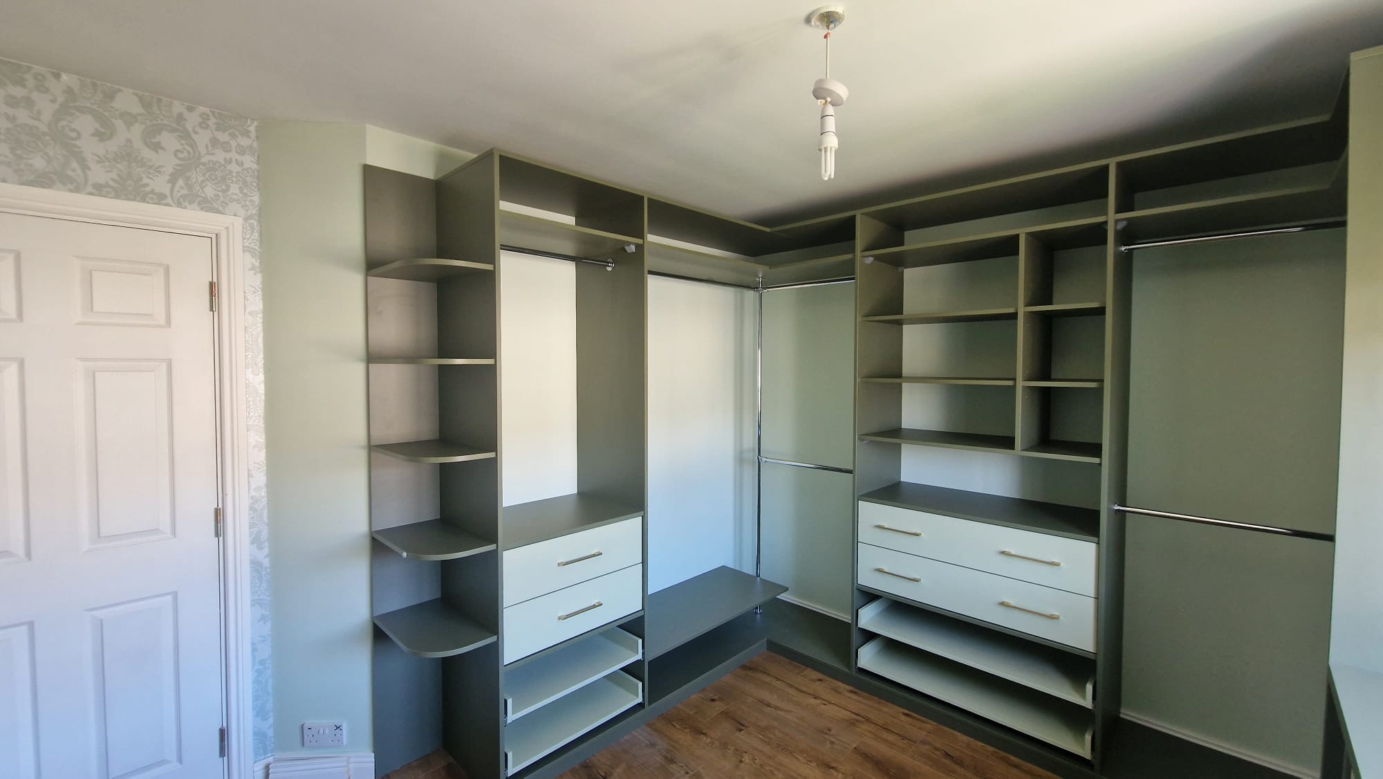 Sage green fitted wardrobes with open shelves, hanging rails, and soft-close drawers. Custom corner bedroom storage with gold handles and wood-effect flooring.