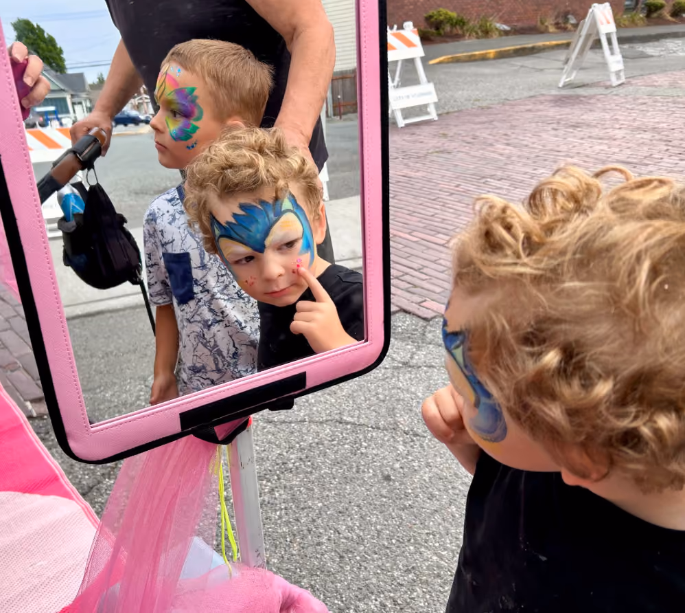 Two young boys with colorful face paint looking into a pink-framed mirror outdoors.