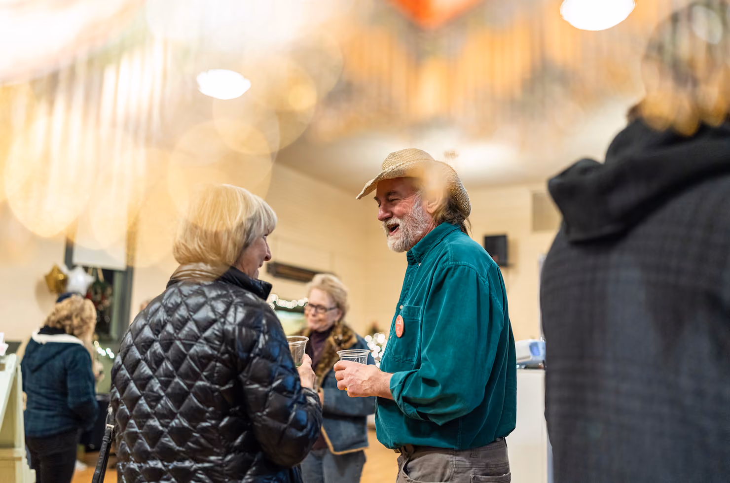 Two older adults smiling and chatting indoors while holding drinks at a social gathering.