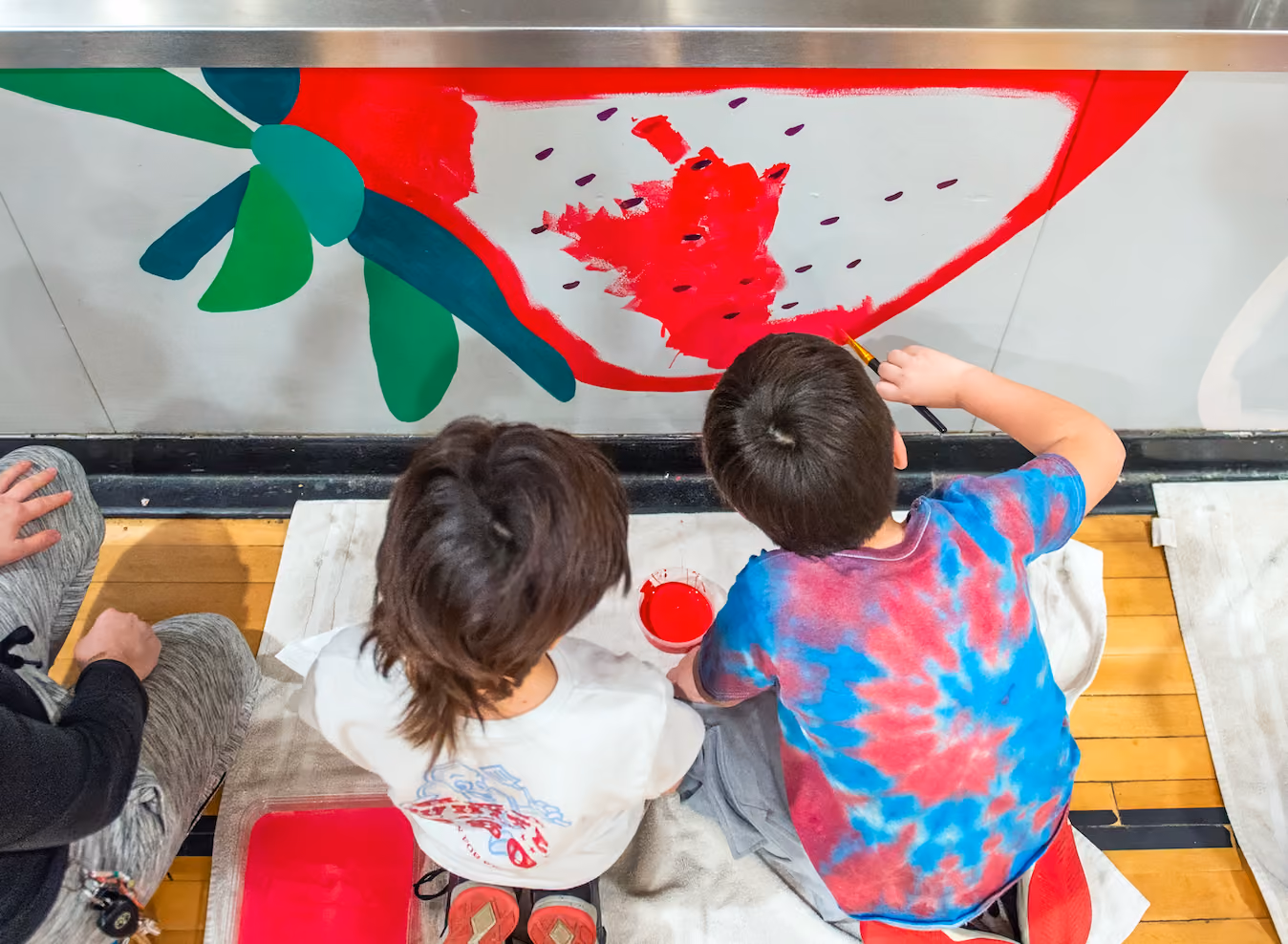 Two children painting a large red watermelon slice mural on a wall indoors.