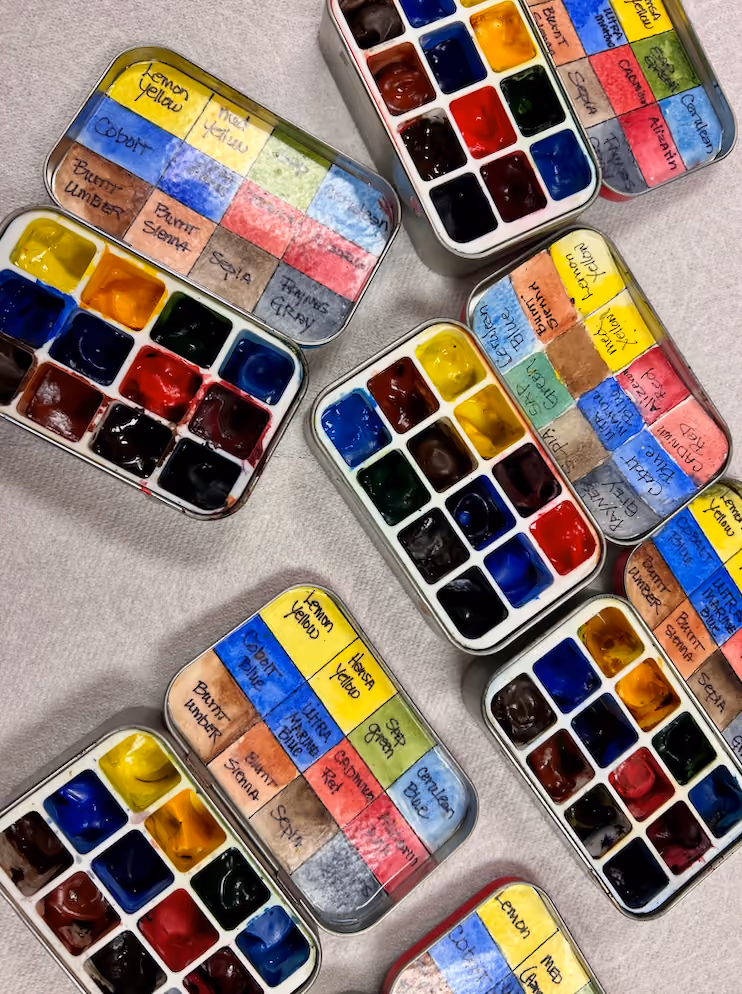 Multiple small watercolor paint palettes with labeled colors arranged on a white cloth.