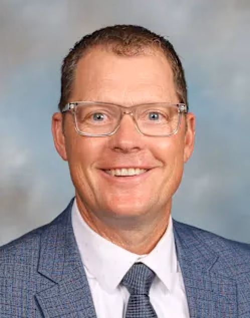 Smiling middle-aged man wearing glasses, a blue patterned suit jacket, white shirt, and gray tie.