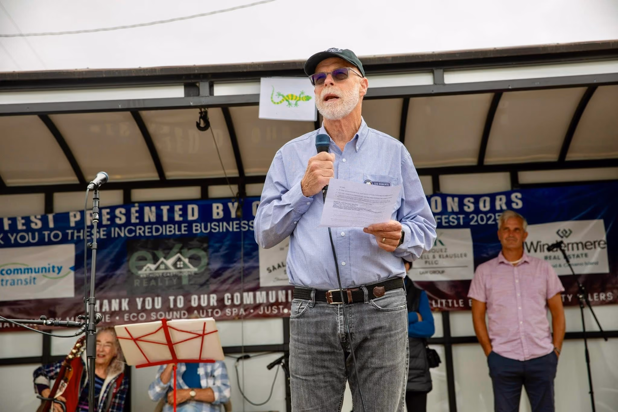 Man in glasses and cap speaking into a microphone onstage with people and sponsor banners behind him.