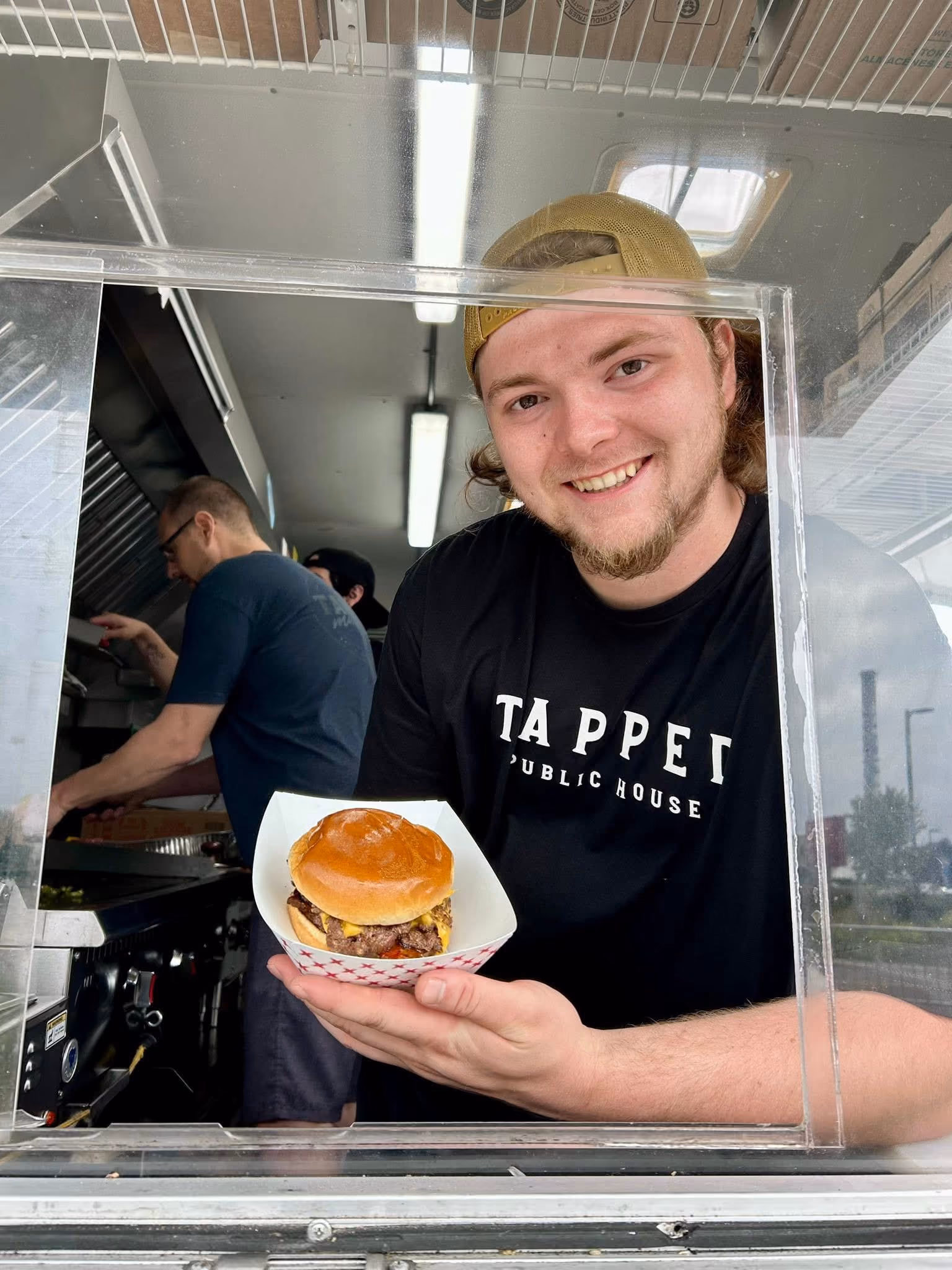 Man in a cap smiles and holds a cheeseburger from a food truck window.