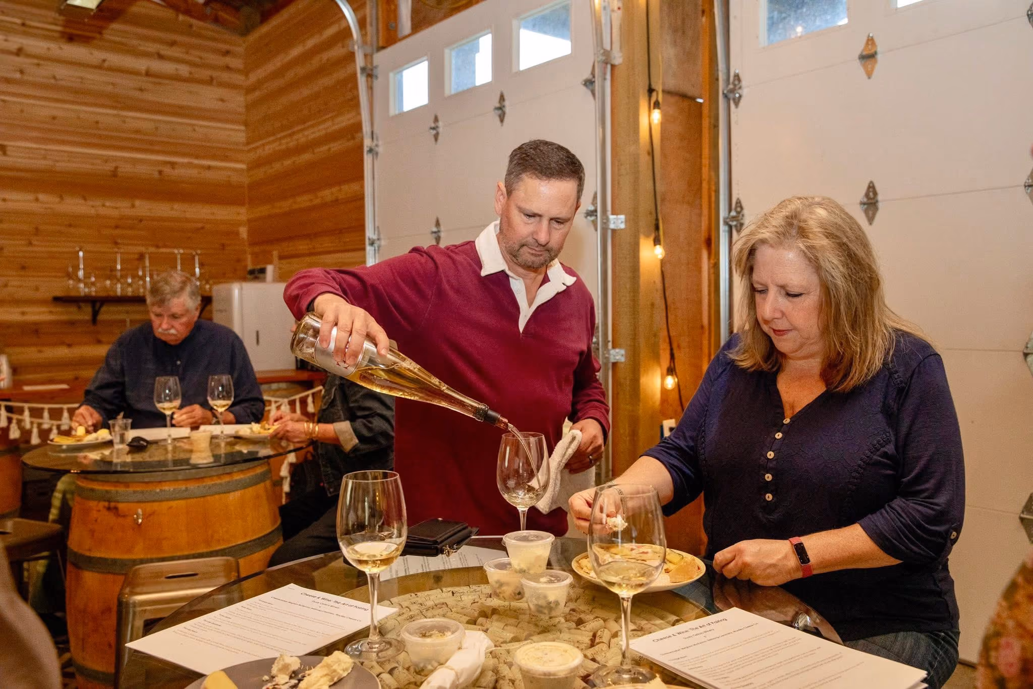 Man pours white wine into glass while woman eats at a table with wine and food in a wooden room.