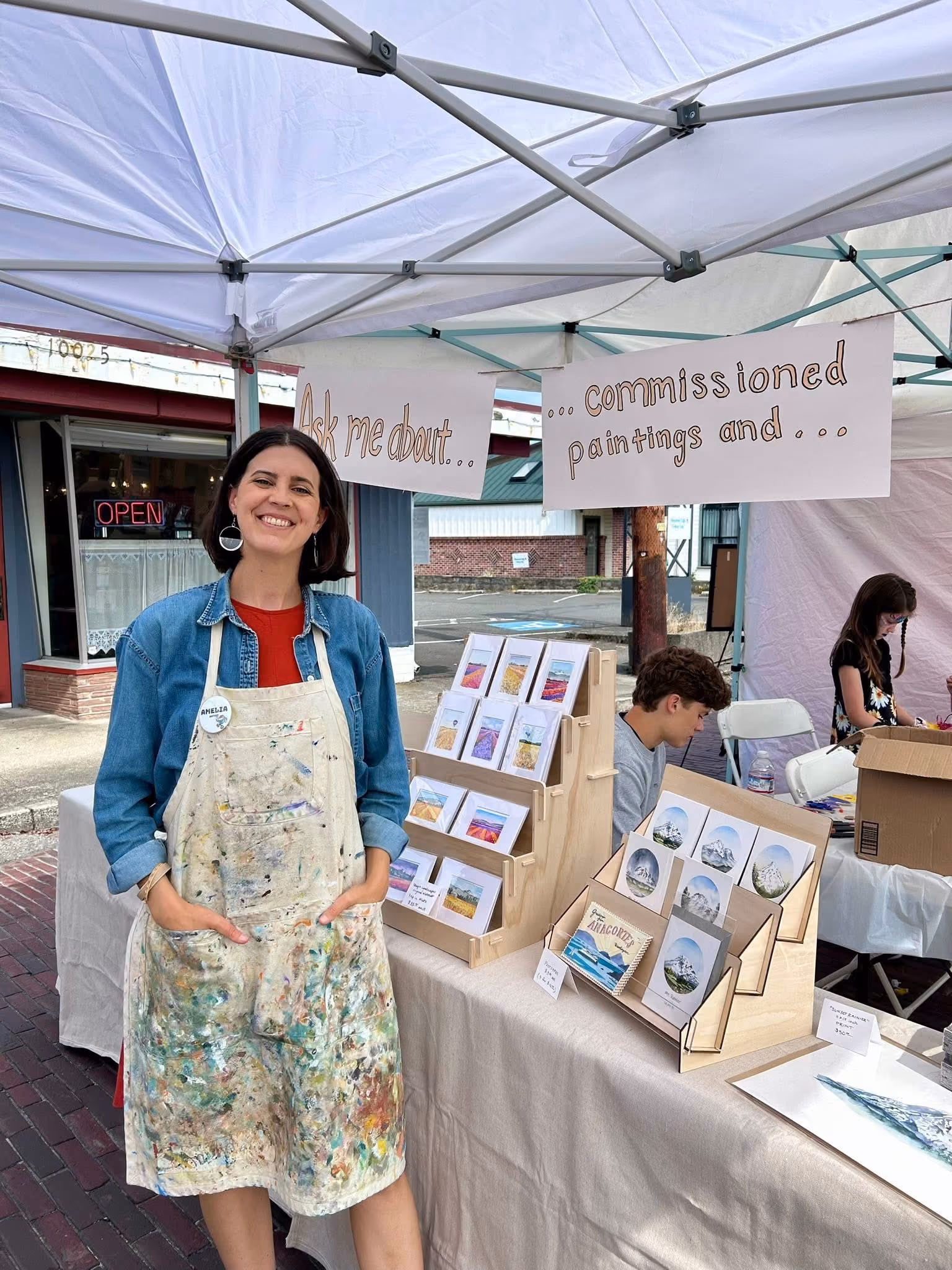 Smiling artist in paint-stained apron standing by table of small landscape artworks at outdoor market.