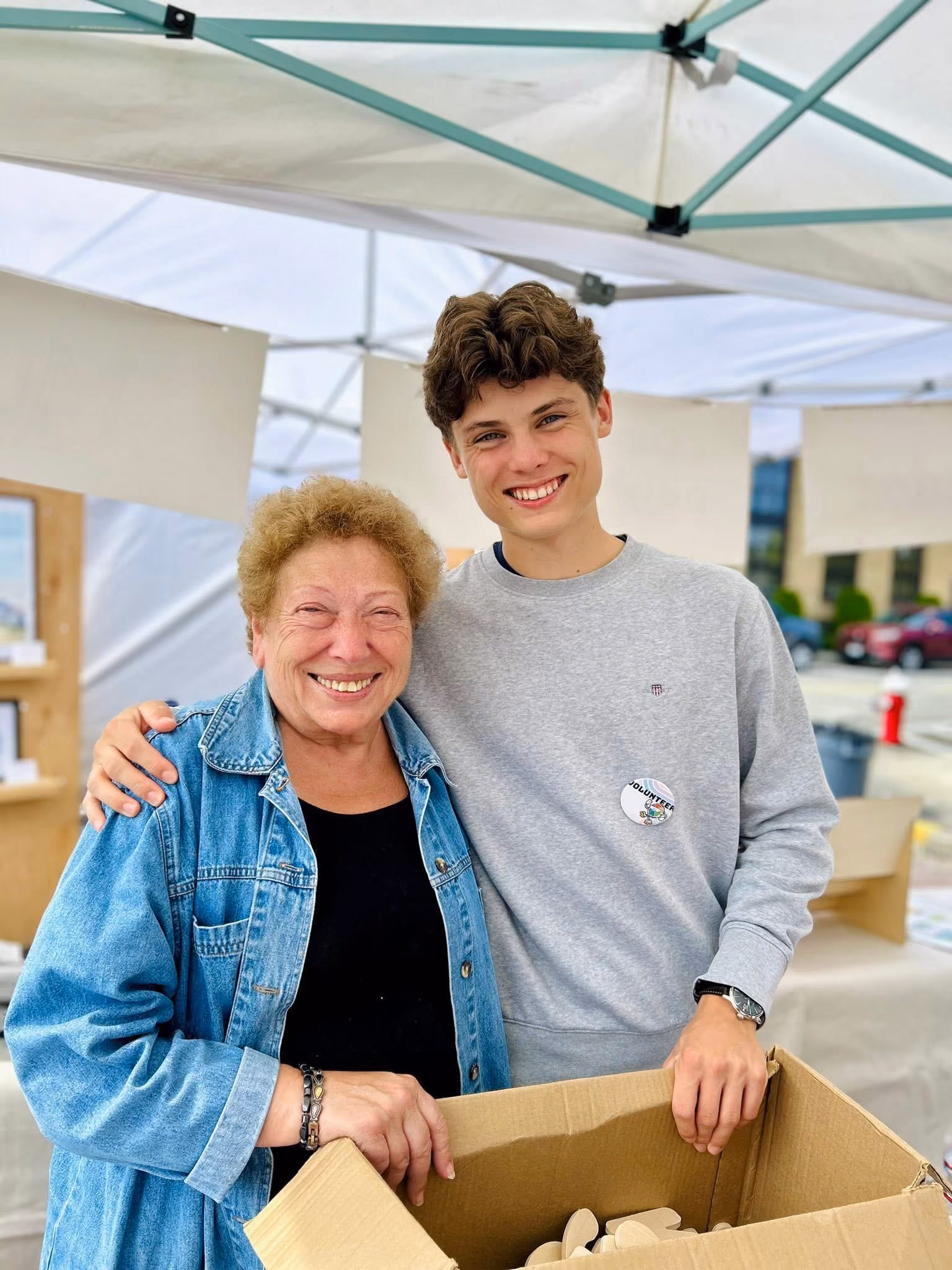 Smiling older woman and young man holding a cardboard box under a white tent at an outdoor event.
